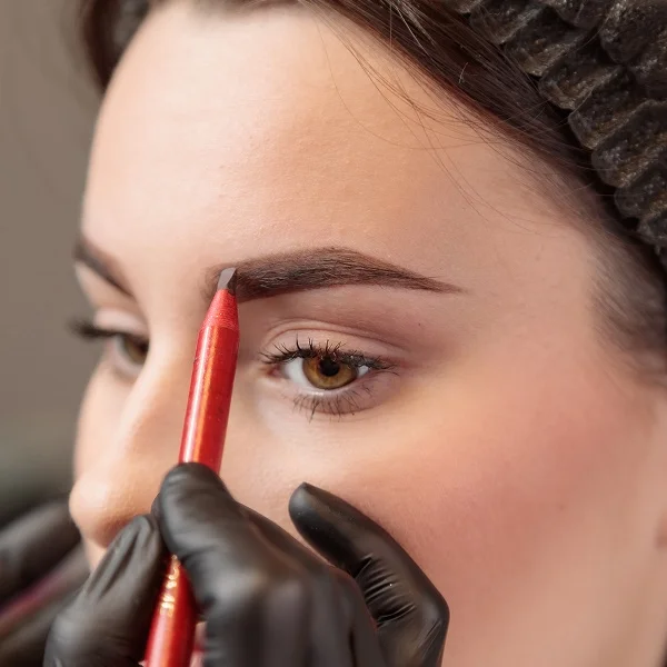 Une esthéticienne portant des gants noirs utilise un crayon de traçage rouge pour définir la forme du sourcil d'une cliente avant une intervention.