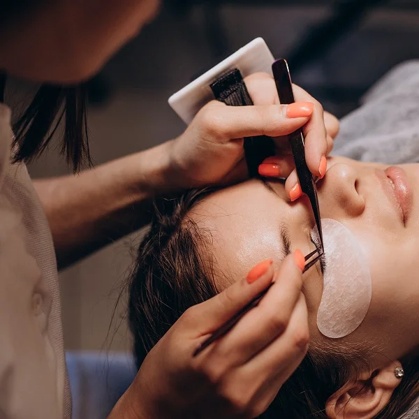 Une technicienne utilise des pinces de précision pour appliquer méticuleusement des extensions de cils sur une cliente allongée.