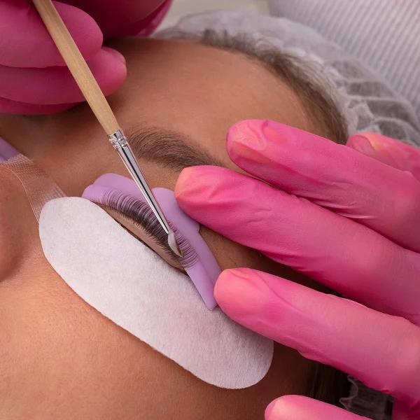 Une esthéticienne portant des gants roses utilise un petit pinceau pour appliquer un soin lors d'un rehaussement de cils sur une cliente.