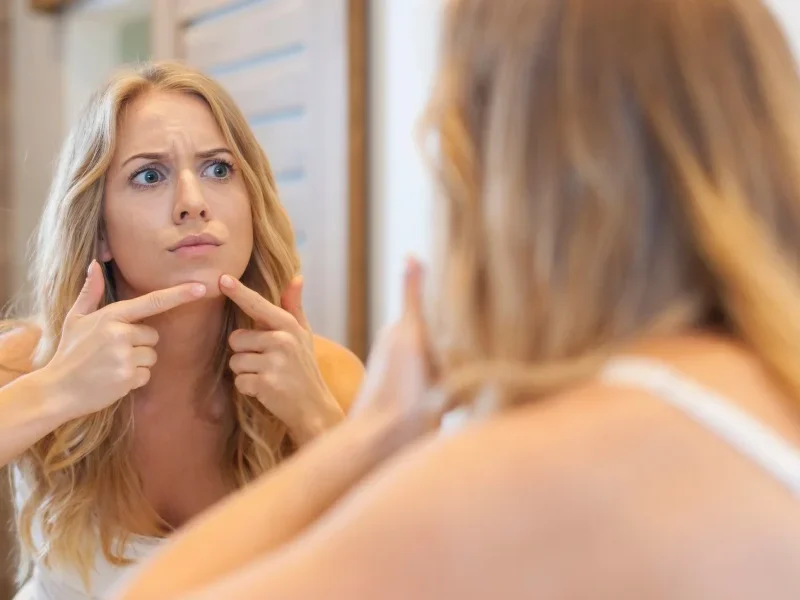A woman gazes into a mirror, examining her reflection and facial features with a thoughtful expression.