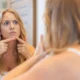 A woman gazes into a mirror, examining her reflection and facial features with a thoughtful expression.