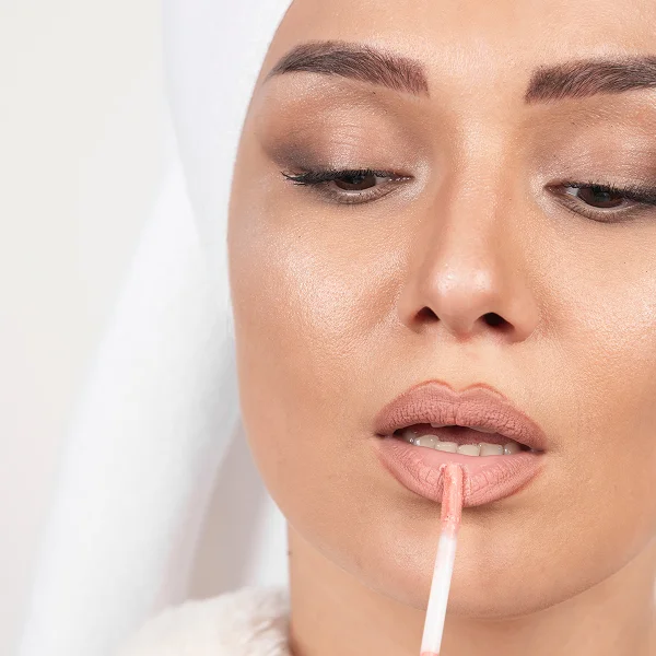 Une femme avec une serviette blanche sur la tête applique un produit repulpeur sur ses lèvres.